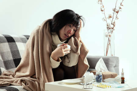 Headache. Young woman wrapped in a plaid looks sick, ill, sneezing and coughing sitting on sofa at...
