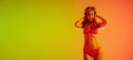 Beautiful seductive girl in fashionable red swimsuit on bright gradient green-orange background in neon light. Full-length portrait. Copyspace for ad. Summer, fashion, beauty, emotions concept. Flyerの写真素材