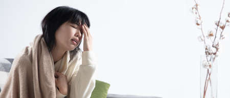Young woman wrapped in a plaid looks sick, ill, sneezing and coughing sitting on sofa at home indoors. Healthcare and medicine, ill prevention, seasonal illness symptoms and self-protection. Flyerの写真素材