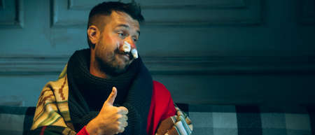 Young man wrapped in a plaid looks sick, ill, sneezing and coughing sitting on sofa at home indoors. Healthcare and medicine, ill prevention, seasonal illness symptoms and self-protection. Flyerの写真素材