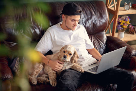 Young man working on laptop computer sitting at home with his cute dog, pet American Cocker Spaniel...