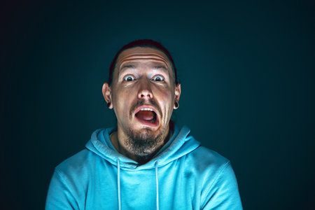 Spooky. Portrait of young crazy scared and shocked caucasian man isolated on dark background. Copyspace for ad. Bright facial expression, human emotions concept. Watching horror on TV, cinema.の写真素材