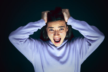 Holding head. Portrait of young crazy scared and shocked caucasian woman isolated on dark...