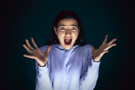 Screaming. Portrait of young crazy scared and shocked caucasian woman isolated on dark background. Copyspace for ad. Bright facial expression, human emotions concept. Looking horror on TV, cinema.の写真素材