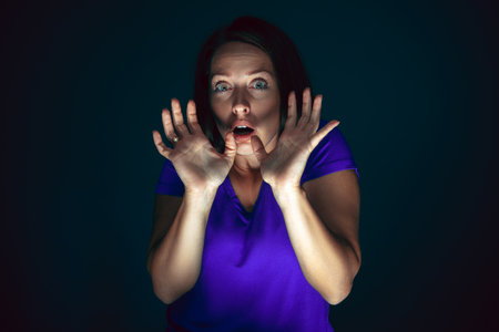 Hiding. Portrait of young crazy scared and shocked caucasian woman isolated on dark background....