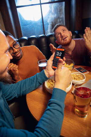 Tensioned. Excited fans in bar with beer and mobile app for betting, score on their devices. Screen...