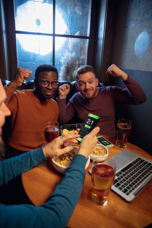 Astonished. Excited fans in bar with beer and mobile app for betting, score on their devices. Screen with match results, emotional friends cheering. Gambling, sport, finance, modern techn concept.の写真素材