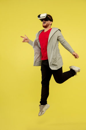 Playing VR-headset. Full length portrait of young high jumping man gesturing isolated on yellow...