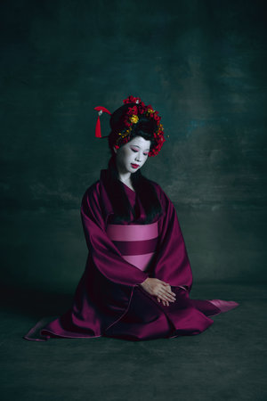 Humble. Young japanese woman as geisha isolated on dark green background. Retro style, comparison of eras concept. Beautiful female model like bright historical character, old-fashioned.の写真素材