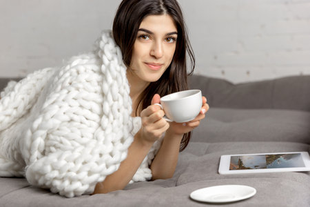 Drinking coffee wrapped in plaid. Portrait of beautiful brunette woman in comfortable soft longsleeve isolated on white studio background. Home comfort, human emotions, winter mood concept. Copyspace.の写真素材