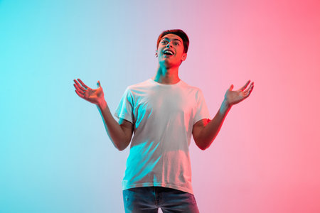 Astonished, greeting. Young caucasian mans portrait on gradient blue-pink studio background in neon light. Concept of youth, human emotions, facial expression, sales, ad. Half length, copyspace.の写真素材