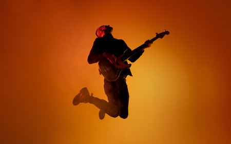 Inspired. Silhouette of young male guitarist isolated on orange gradient studio background in neon light. Beautiful shadow in action, performing. Concept of human emotions, expression, ad, music, art.の写真素材