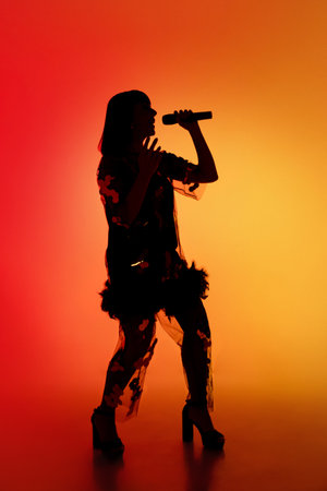 Talented. Silhouette of young female singer isolated on orange gradient studio background in neon light. Beautiful shadow in action, performing. Concept of human emotions, expression, ad, music, art.の写真素材