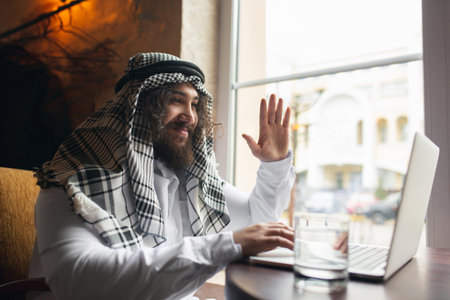 Videocall. Arabian businessman working in office, business centre using device, gadget. Modern saudi lifestyle. Man in traditional wear and scarf looks confident, busy, handsome. Ethnicity, finance.の写真素材