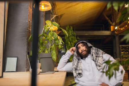 On the phone. Arabian businessman working in office, business centre using device, gadget. Modern saudi lifestyle. Man in traditional wear and scarf looks confident, busy, handsome. Ethnicity, finance.の写真素材