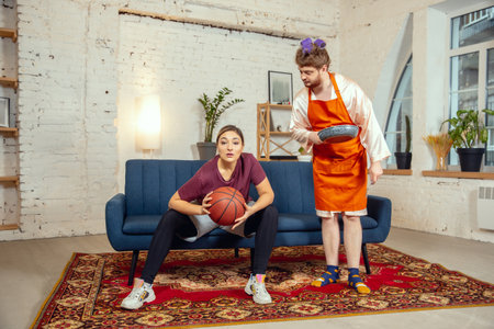 Gender stereotypes. Wife and husband doing things unusual for their genders in social meanings, sense. Man cooking dinner while woman training in basketball with the ball in living room.の写真素材