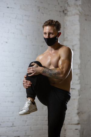 Stretching. Professional male athlete training on brick wall background wearing face mask. Sport during quarantine of coronavirus worldwide pandemic. Young man practicing in gym safe using equipment.の写真素材