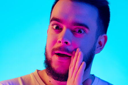 Secrets. Caucasian mans portrait isolated on blue studio background in neon light. Beautiful male model. Concept of human emotions, facial expression, sales, ad. Copyspace for ad. Close upの写真素材