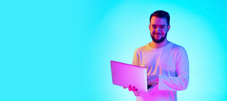 Using laptop. Caucasian mans portrait with device on blue studio background in neon light. Beautiful male model. Concept of human emotions, facial expression, sales, ad. Copyspace for ad, flyerの写真素材