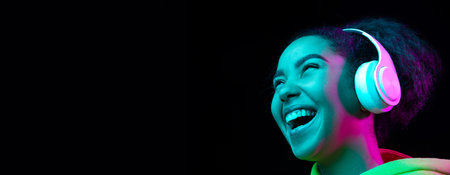 Laughting, flyer. African-american woman isolated on dark background in multicolored neon. Listening to music with headphones. Concept of human emotions, facial expression, sales, ad, fashion.の写真素材