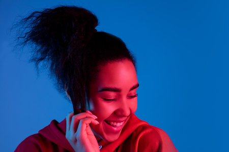 Cute and shy. African-american womans portrait on blue studio background in red-pink neon light. Beautiful female model. Concept of human emotions, facial expression, sales, ad, fashion. Copyspace.の写真素材