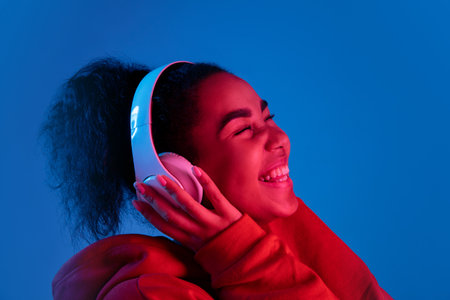 Happiness. African-american womans portrait on blue background in red-pink neon light. Listening to...