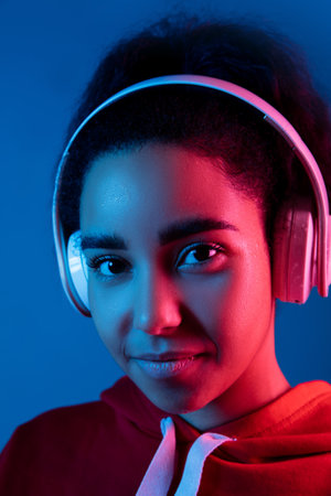 Attented. African-american womans portrait on blue background in red-pink neon light. Listening to music with headphones. Concept of human emotions, facial expression, sales, ad, fashion. Copyspace.の写真素材
