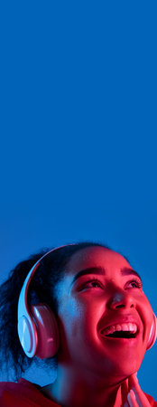 Flyer. African-american womans portrait on blue background in red-pink neon light. Listening to music with headphones. Concept of human emotions, facial expression, sales, ad, fashion. Copyspace.の写真素材