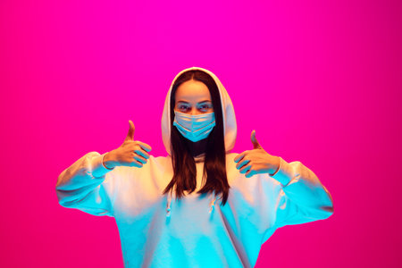 Thumbs up. Caucasian womans portrait on pink studio background in mixed neon light. Using protective face mask. Concept of human emotions, facial expression, coronavirus, healthcare. Copyspace.の写真素材