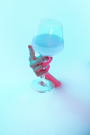 Fashionable. Close up female hand gesturing from the milk bath with soft white glowing in neon light. Copyspace for advertising. Holding glass of blue cocktail, alcohol. Modern neoned colors.の写真素材