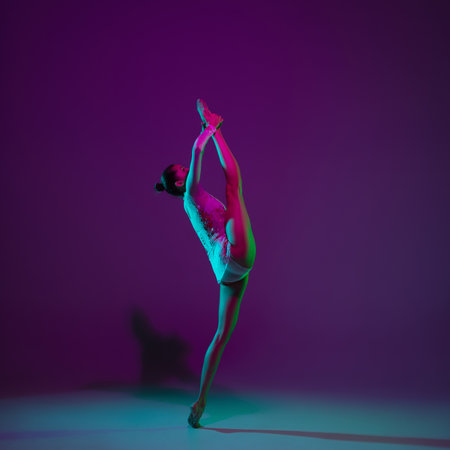 Motion. Young female athlete, rhythmic gymnastics artist dancing, training isolated on purple studio background in neon light. Beautiful girl practicing with equipment. Grace in performance.の写真素材