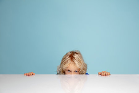 Hiding. Happy curly boy isolated on blue studio background. Looks happy, cheerful. Copyspace for ad. Childhood, education, emotions, facial expression concept. Peeking out from behind the tableの写真素材