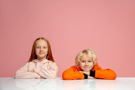 Excellent. Happy children isolated on coral pink studio background. Look happy, cheerful. Copyspace for ad. Childhood, education, emotions, facial expression concept. Sitting at the table dreamful.の写真素材