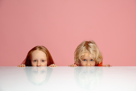 Playful. Happy children isolated on coral pink studio background. Look happy, cheerful. Copyspace for ad. Childhood, education, emotions, facial expression concept. Peeking out from behind the table.の写真素材