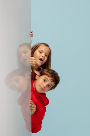 Looking up. Happy children isolated on blue studio background. Look happy, cheerful. Copyspace for ad. Childhood, education, emotions, facial expression concept. Peeking out from behind the wall.の写真素材