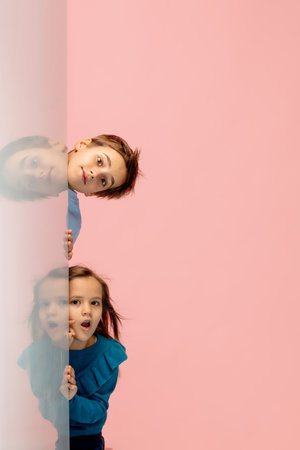 Surprised. Happy children isolated on coral pink studio background. Look happy, cheerful. Copyspace for ad. Childhood, education, emotions, facial expression concept. Peeking out from behind the wall.の写真素材