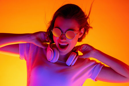 Drive. Caucasian womans portrait on orange studio background in red-pink neon light. Beautiful female model in sunglasses and device. Concept of human emotions, facial expression, sales, ad, fashion.の写真素材