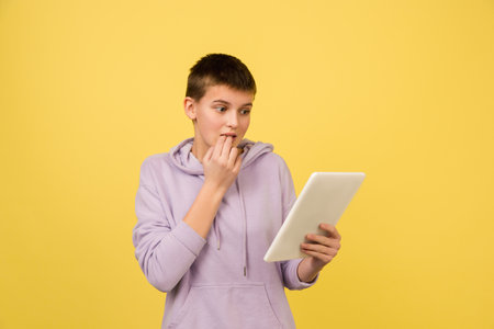 Scared scrolling tablet. Caucasian girls portrait isolated on yellow background with copyspace. Beautiful female model in hoodie. Concept of human emotions, facial expression, sales, ad, fashion.の写真素材
