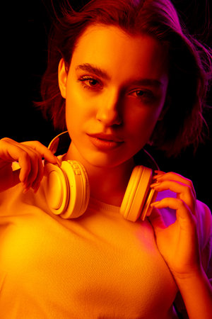 Style. Caucasian womans portrait on black studio background in pink-orange neon light. Beautiful female model with headphones. Concept of human emotions, facial expression, sales, ad, fashion.の写真素材