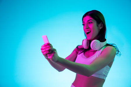 Chatting. Brunette womans portrait on blue studio background in mixed neon. Beautiful model with headphones and phone. Concept of human emotions, facial expression, sales, ad, fashion, devices.の写真素材