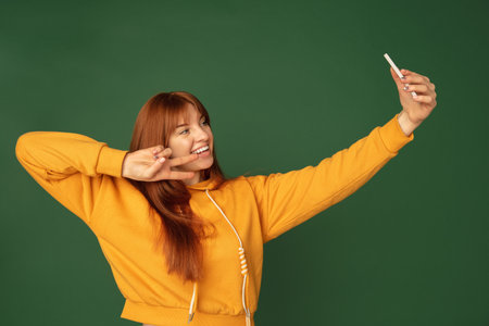 Selfie, smiling. Caucasian womans portrait isolated on green studio background with copyspace. Beautiful female model with phone. Concept of human emotions, facial expression, sales, ad, fashion.の写真素材