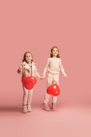 Hearts. Happy kids, girls isolated on coral pink studio background. Look happy, cheerful. Copyspace for ad. Childhood, education, emotions, facial expression concept. Jumping, running having funの写真素材