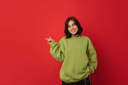 Smiling, pointing. Caucasian womans portrait isolated on red background with copyspace. Beautiful female model in green hoodie. Concept of human emotions, facial expression, sales, ad, fashion.の写真素材