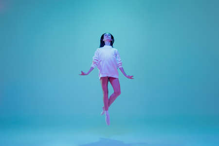In flight. Young and graceful ballet dancer isolated on blue studio background in neon light. Art, motion, action, flexibility, inspiration concept. Flexible caucasian ballet dancer, moves in glow.の写真素材