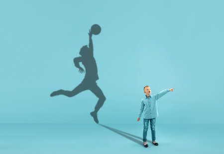 Childhood and dream about big and famous future. Conceptual image with boy and shadow of sportive male basketball player, champion on blue background. Childhood, dreams, imagination, education concept.の写真素材