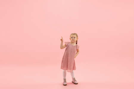 Pointing. Childhood and dream about big and famous future. Pretty longhair girl on coral pink studio background. Childhood, dreams, imagination, education, facial expression, emotions concept.の写真素材