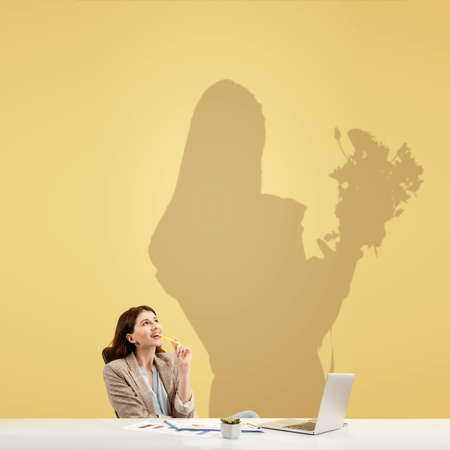 Young caucasian woman dreaming about future in floristics during her work in office. Shadow, silhouette of female florist on the wall behind her. Becoming a legend. Inspiration, aspiration. Copyspace.の写真素材