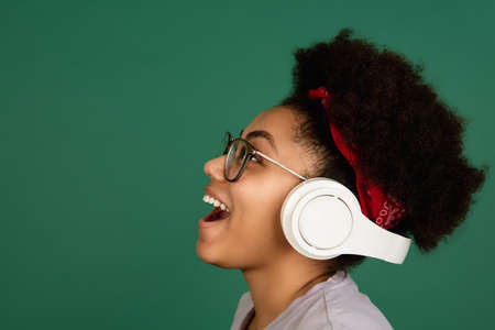 Music. African-american beautiful womans portrait isolated on green studio background with copyspace. Female model with headphones. Concept of human emotions, facial expression, sales, ad, fashion.の写真素材