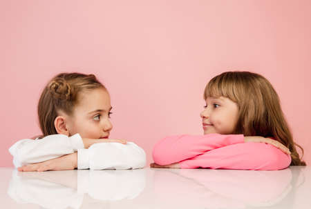 Kindly. Happy kids, girls isolated on coral pink studio background. Look happy, cheerful. Copyspace for ad. Childhood, education, emotions, business, facial expression concept. Sitting at the tableの写真素材