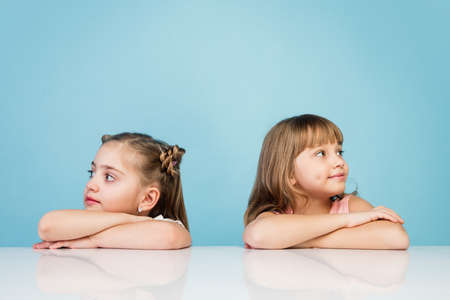 Happy kids, girls isolated on blue studio background. Look happy, cheerful. Copyspace for ad. Childhood, education, emotions, business, facial expression concept. Sitting at the table, dreamingの写真素材
